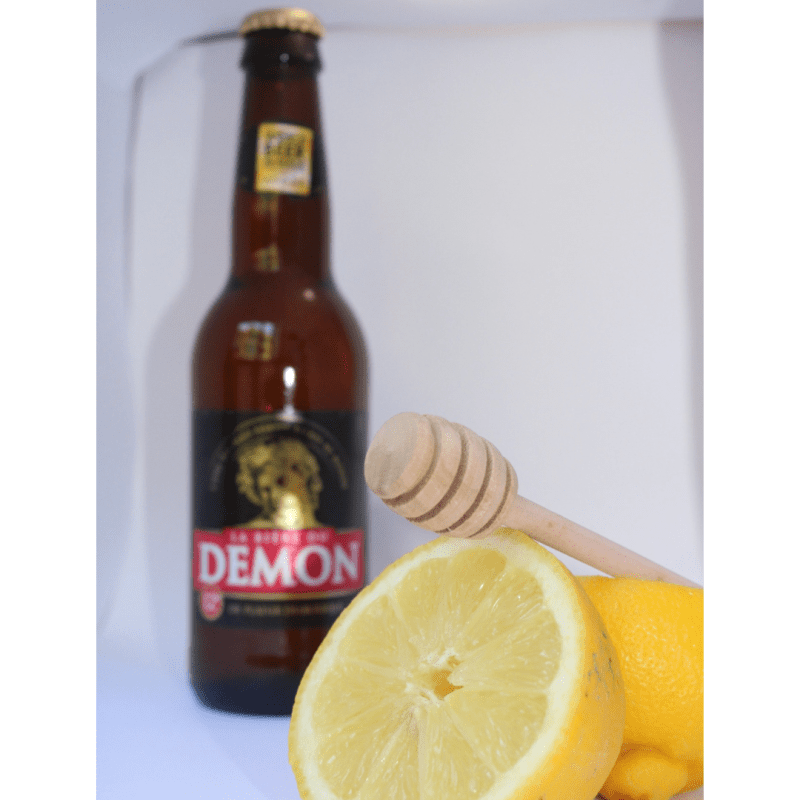 Botella de La Bière du Démon con limón fresco y palo mielero sobre fondo blanco, destacando los aromas cítricos y dulces de esta cerveza francesa de 12% ABV