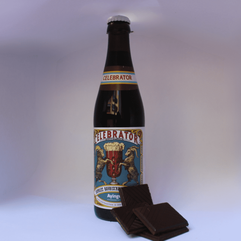 Botella de cerveza Ayinger Celebrator Doppelbock con pastillas de chocolate negro, sobre fondo blanco, ideal para destacar su maridaje y notas de sabor tostadas y dulces.